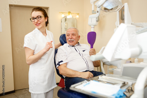 Smiling and happy senior man 70-75 years old on review of young happy woman dentist, sitting in dental chair in dental office. Dental care for elderly. Dentistry, medicine and healthcare concept
