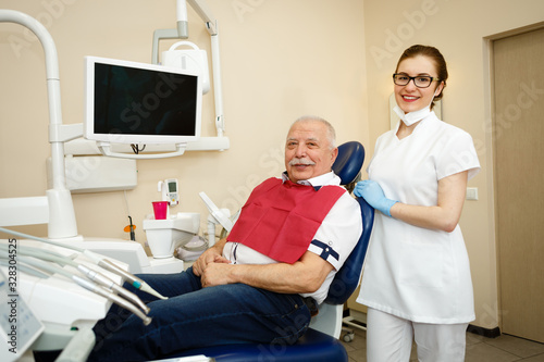Portrait of smiling senior man with mustache 70-75 years old sitting in dental chair after teeth treatment and young woman dentis looking at camera. Concept of dentistry, medicine and health care