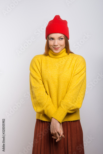Shy smiling woman in stylish knitwear. Beautiful happy young woman in red knitted hat and yellow sweater smiling and looking aside isolated on white background. Style and emotion concept © Mangostar