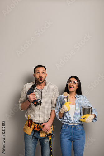 excited manual workers holding paint can, brushes and electric drill on grey
