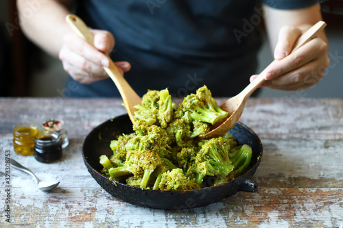 A chef with a wooden spatula and spoon mixes broccoli in a pan.