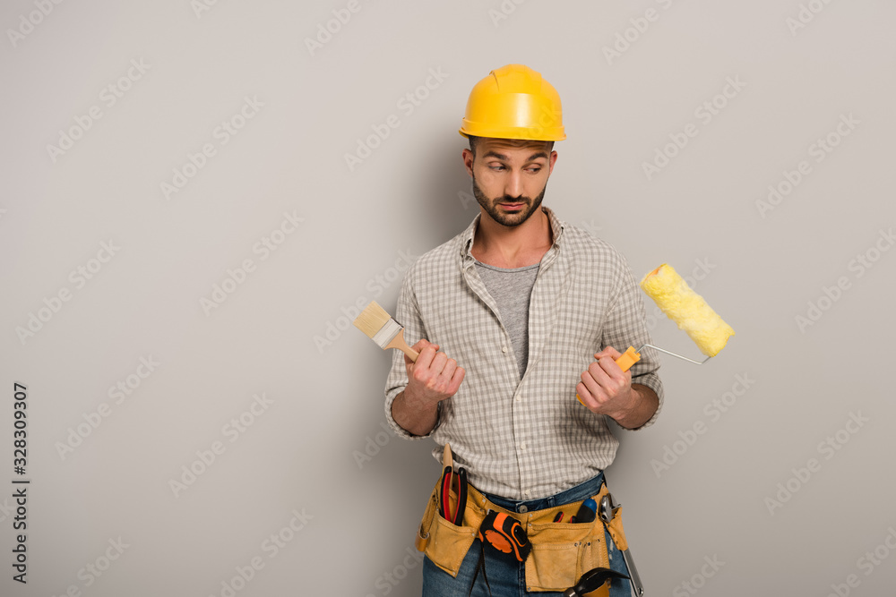 skeptical manual worker in hardhat holding paint roller and brush on grey skeptical manual worker in hardhat holding paint roller and brush on grey