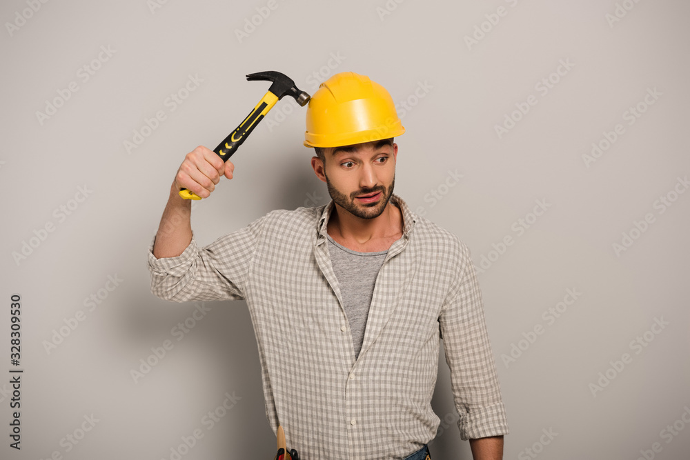 thoughtful repairman in helmet holding hammer on grey thoughtful repairman in helmet holding hammer on grey