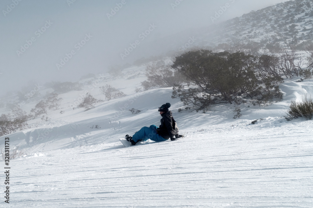 Snowboarder sitting facing foggy hill.