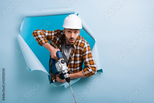 surprised manual worker in helmet holding electric drill in torn paper, isolated on blue