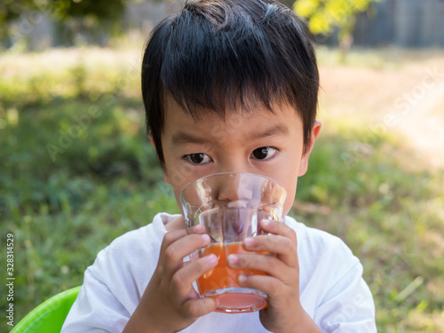 Asian little child boy drinking orange juice from glass outdoor nature background at home. Vitamin C nutrition for healthy concept.