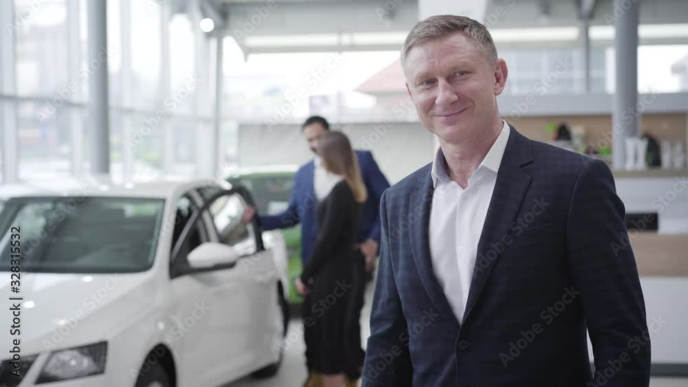 Confident Caucasian car dealer looking back at happy multiracial couple opening new white car and showing keys at camera. Smiling man posing in dealership. Showroom. business, lifestyle. Middle shot. Confident Caucasian car dealer looking back at happy multiracial couple opening new white car and showing keys at camera. Smiling man posing in dealership. Showroom. business, lifestyle. Middle shot.