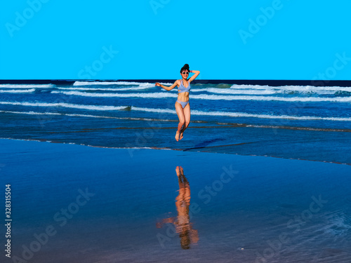 Happy young woman with a beautiful body wearing bikinis showing abs and jumping on beach shore - Blue highlights and shadows - Summer Trip Concept