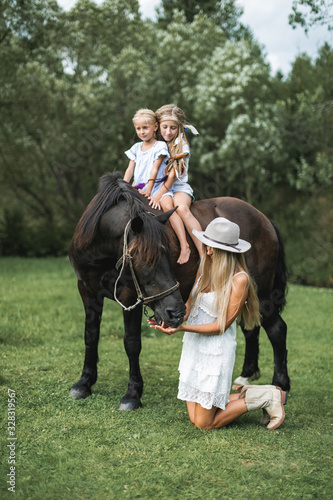 Happy family, mother with two children cute daughters riding a horse. Mother in white dress, hat and cowboy boots standing on the knees and feeding the horse. Family time on nature, horse riding
