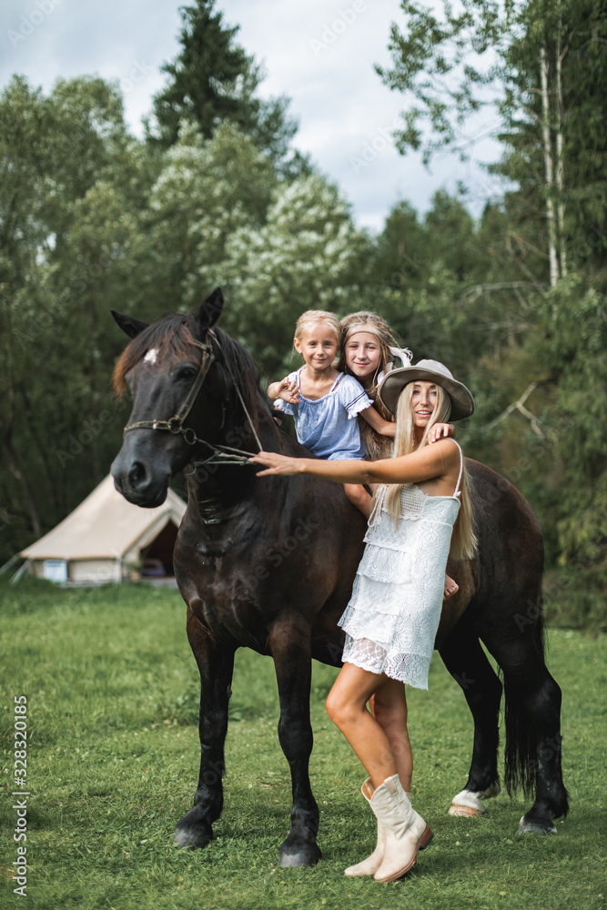 Young beautiful mother in white dress, hat and cowboy boots, petting horse, while her two cute little daughters riding the horse in countryside. Wigwam tent on the background Young beautiful mother in white dress, hat and cowboy boots, petting horse, while her two cute little daughters riding the horse in countryside. Wigwam tent on the background