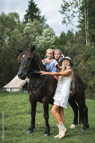 Young beautiful mother in white dress, hat and cowboy boots, petting horse, while her two cute little daughters riding the horse in countryside. Wigwam tent on the background