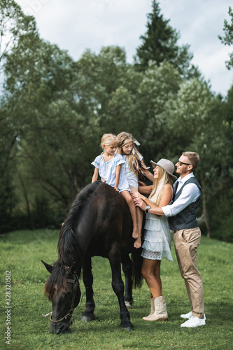 Young stylish family in cowboy wear, father, mother teaching their two little daughters to ride a horse. Family horse riding on the nature in countryside © sofiko14