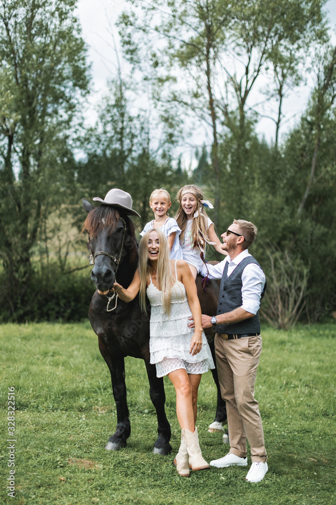 Happy family, parents and children, enjoying the horse presence. Girls are riding the horse, wearing hat. Man and woman parents having fun and laughing. Countryside, horse riding outdoors