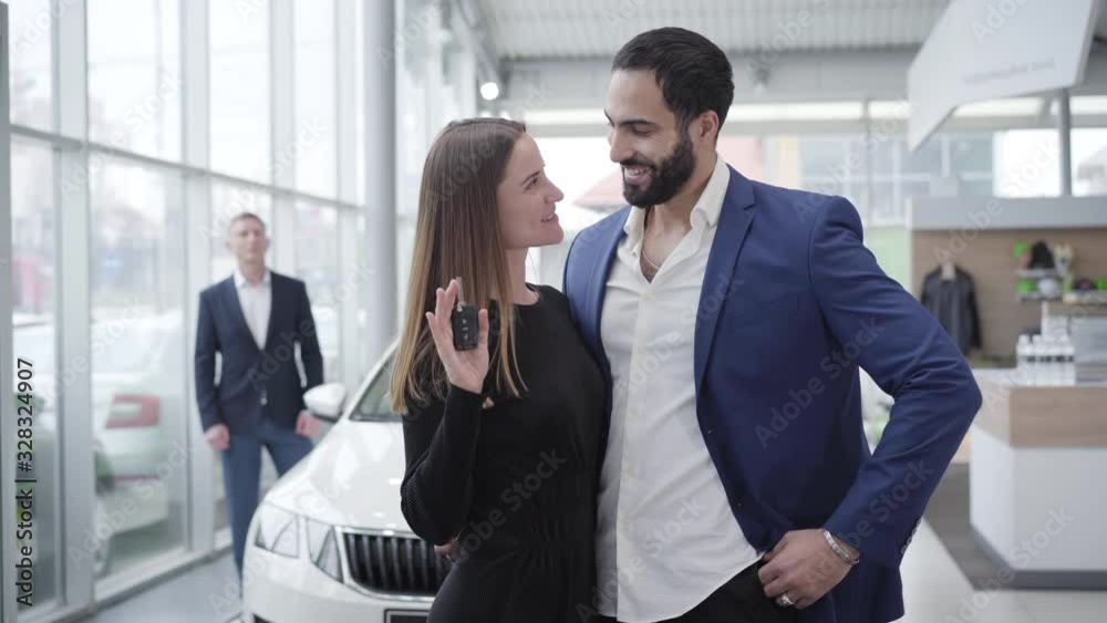 Happy young Caucasian woman talking car keys from Middle Eastern man in dealership and smiling at camera. Blurred trader opening new white vehicle at the background. Success, luxury, lifestyle. Happy young Caucasian woman talking car keys from Middle Eastern man in dealership and smiling at camera. Blurred trader opening new white vehicle at the background. Success, luxury, lifestyle.