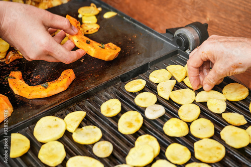 Orange butternut squash pieces grilled on electric grill, detail on woman hands moving smoking vegetables, blurred potato chips in front