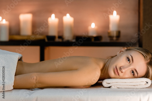 Charming young woman lies on stomach with beauty smile on massage table against background of blurry candles waiting for massage. Healthy lifestyle and relaxation concept