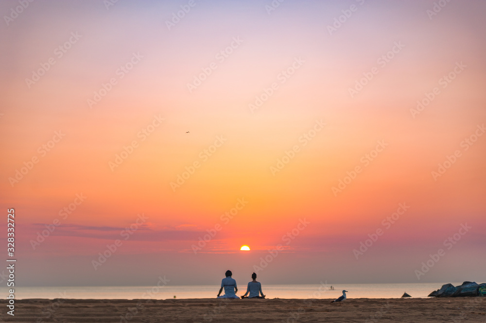 Fototapeta premium couple doing a relaxing yoga and meditation session on the beach at sunrise with birds and the sun rising