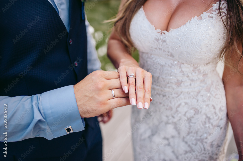 The groom in a suit and the bride in a white dress stand on the background of a green park and hold their hands together, there are wedding rings on their fingers