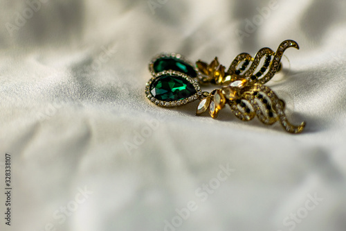 Metal gold earrings with green, emerald stones, white and yellow rhinestones. On a white fabric flattened background. Brilliant and festive decoration
