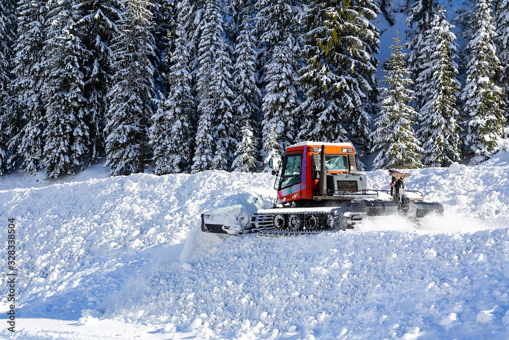 A snow removal and ski tracks making machine removes snow in a ski resort in winter. Snow-covered Forest on the hill is in the background. A snow removal and ski tracks making machine removes snow in a ski resort in winter. Snow-covered Forest on the hill is in the background.