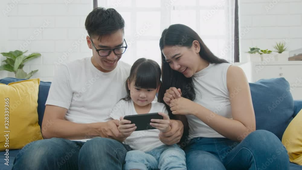 Slow motion zooming shot : Happy warm family father, mother and daughter playing funny game online on smartphone mobile sitting on couch together, parents with kids enjoying mobile apps on smart phone Slow motion zooming shot : Happy warm family father, mother and daughter playing funny game online on smartphone mobile sitting on couch together, parents with kids enjoying mobile apps on smart phone
