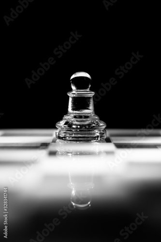Single clear glass pawn chess piece or figure standing on glass chessboard with reflection. Isolated on black background with copy space. Black and white photography.