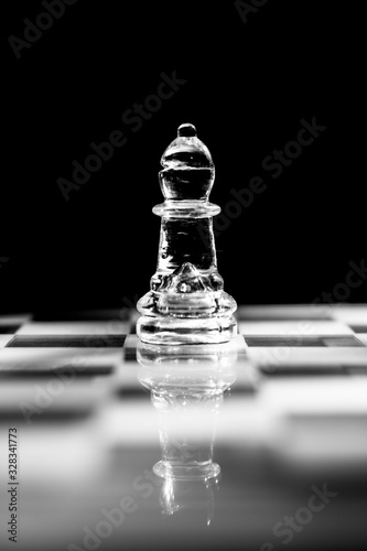 Single clear glass bishop chess piece or figure standing on glass chessboard with reflection. Isolated on black background with copy space. Black and white photography.