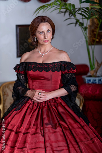 A woman  elegantly dressed  in 19th-century-style clothes  poses standing in the hall of a palace