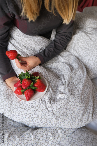 Woman holds a plate with fresh strawberries in bed. Romantic wake up with berries. Relaxation and enjoyment. Cozy and comfortable morning. Healthy organic sweet fruit. Vitamins diet for girls