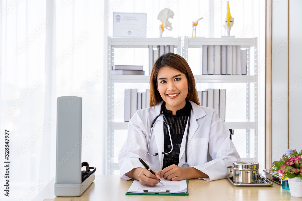 Portrait of a smiling Asian medical doctor sitting at desk in office clinic hospital wearing white coat with stethoscope, looking at camera Portrait of a smiling Asian medical doctor sitting at desk in office clinic hospital wearing white coat with stethoscope, looking at camera