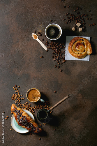 Variety of traditional french puff pastry raisin and chocolate buns, croissant with various cups of coffee and milk, cezve, recycled wooden spoon of sugar over dark texture background. Flat lay, space