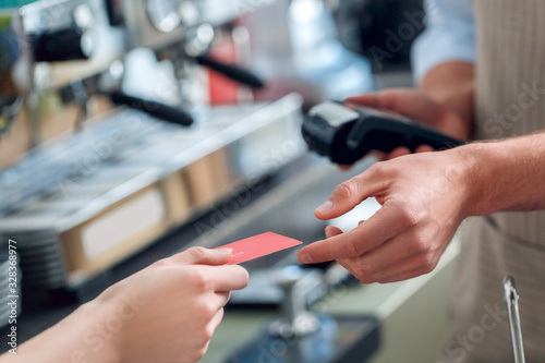Young adult barista holding wireless terminal and taking card from client