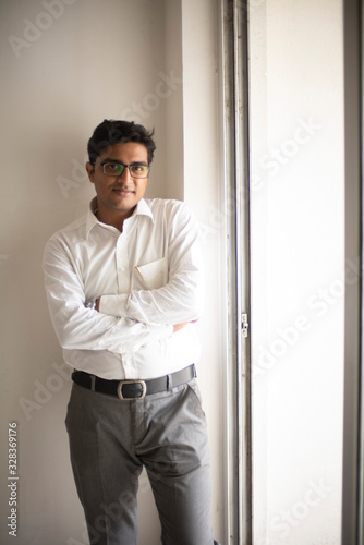 Portrait of an Indian Bengali tall, dark, handsome brunette young man in office wear is standing in front of a glass window in a corporate office/bpo/call center. Indian corporate lifestyle