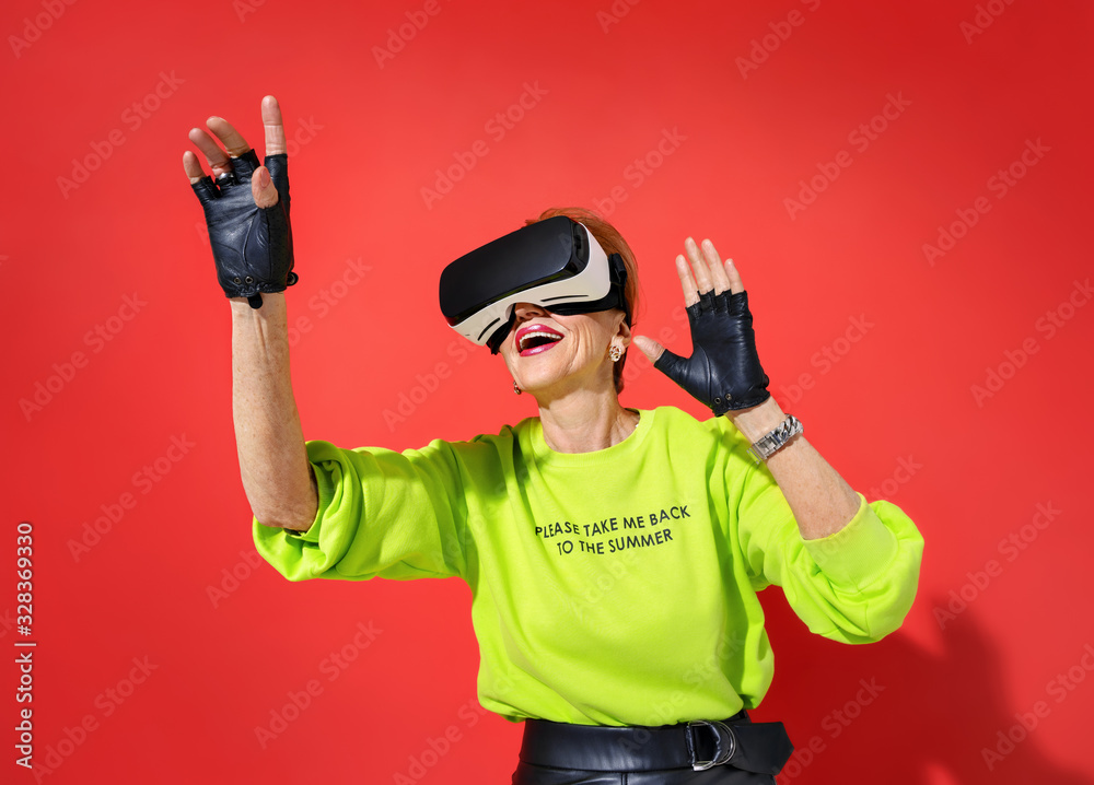 Forever Young! Elderly woman in bright outfit with glasses of virtual reality, touching something on red background