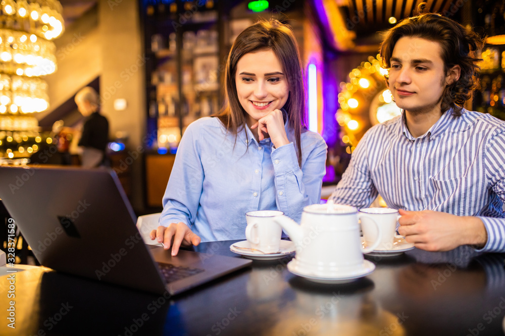 Beautiful young couple is using a laptop, talking and smiling while sitting in the cafe.