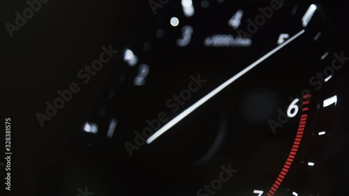 Car dashboard. Black, white numbers and arrows. Night, interior lighting. Tachometer, speedometer, sharp, fast acceleration of the car forward. The arrow rises quickly to the red zone, cutoff