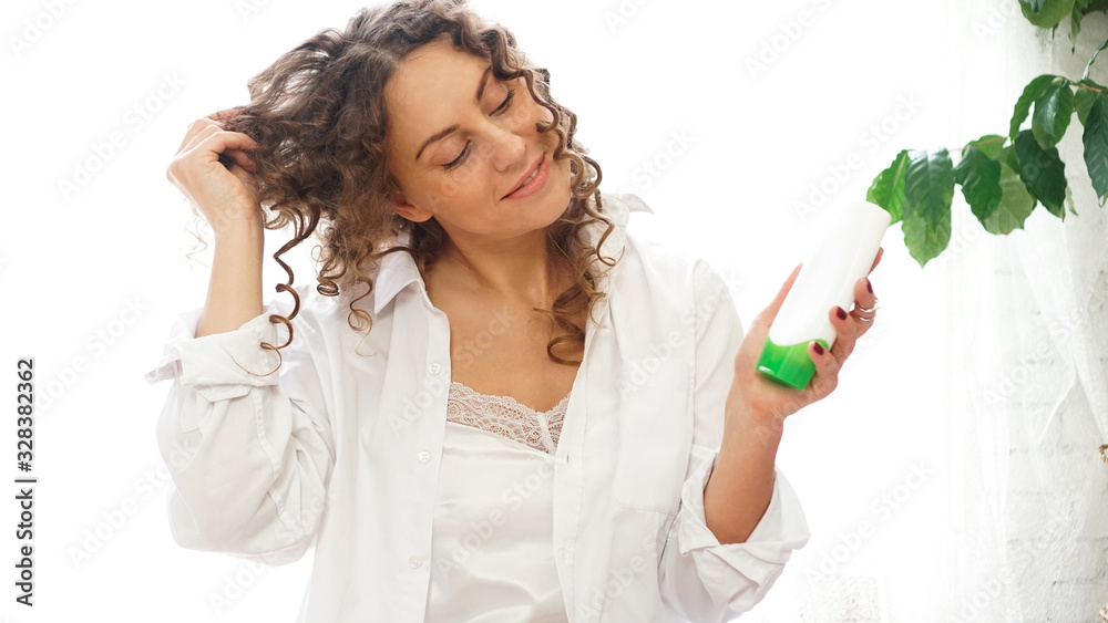 Pretty woman showing white cosmetic tube for curly hair care in her hand at home, white room with green plants Pretty woman showing white cosmetic tube for curly hair care in her hand at home, white room with green plants