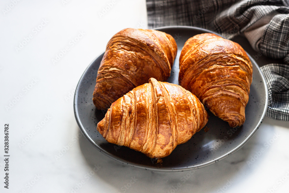 Three delicious fresh croissants on a ceramic plate, light grey background. 