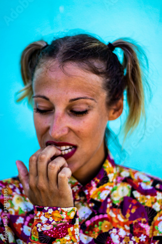 Close up portrait of a young beautiful blond woman with pigtails and hipster style, biting her nails, concentrated expression, colorful Japanese shirt, red lips and blue street wall in the background