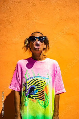Portrait of a cute and funny young woman with pigtails and hipster style sunglasses makes a bubble with her chewing gum, red lips and orange street wall in the background, fashion urban concept