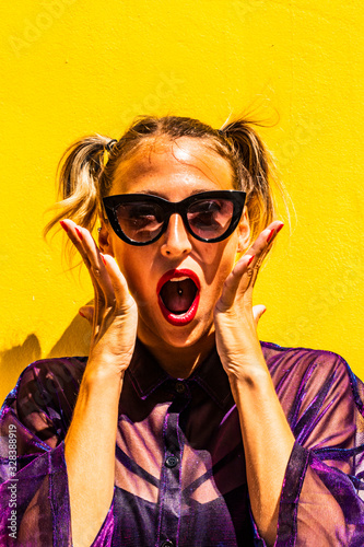 Portrait young woman pigtails and expression of surprise, red lips hipster style, sunglasses and yellow street wall in background, fashion vertical urban concept