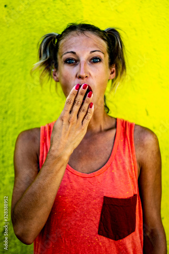 Urban portrait of a cute and funny blond woman in hipster style with pigtails making surprised expression, red lips and neon yellow street wall in the background, fashion vertical concept