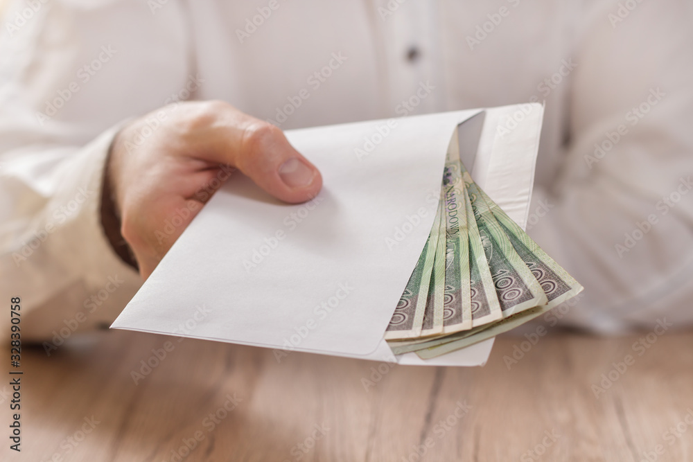 Męska dłoń trzyma białą kopertę wypełnioną banknotami polskiej waluty i podaje ją ponad stołem. - obrazy, fototapety, plakaty 