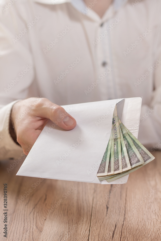 Męska dłoń trzyma białą kopertę wypełnioną banknotami polskiej waluty i podaje ją ponad stołem.