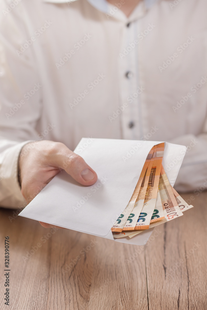 Męska dłoń trzyma białą kopertę wypełnioną banknotami waluty Euro i podaje ją ponad stołem.