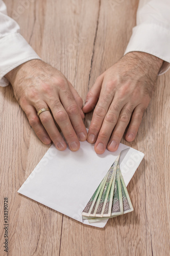 Mężczyzna w białej koszuli trzyma końcami palców białą kopertę wypełnioną banknotami polskiej walucie PLN i przesuwa ją po stole dając łapówkę. 