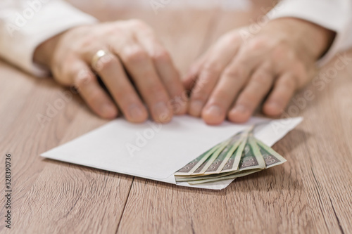 Pieniądze, banknoty w polskiej walucie polski złoty leżą na stole w kopercie. Na drugim planie męskie dłonie trzymają palcami róg koperty.