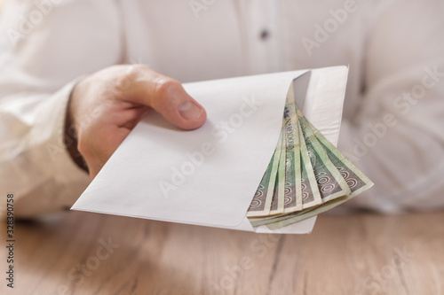 Męska dłoń trzyma białą kopertę wypełnioną banknotami polskiej waluty i podaje ją ponad stołem.