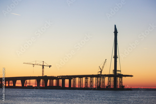 An unfinished part of highway road or bridge, on sunset or dusk, Industrial construction cranes and building silhouettes at sunrise, back light construction site silhouetted at sunset, over the water © tsuguliev