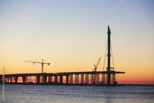 An unfinished part of highway road or bridge, on sunset or dusk, Industrial construction cranes and building silhouettes at sunrise, back light construction site silhouetted at sunset, over the water